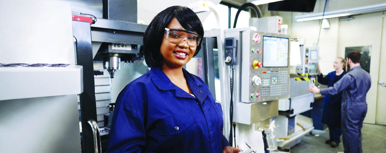 Smiling female mechanical Engineering student poses in front of machine holding tool.
