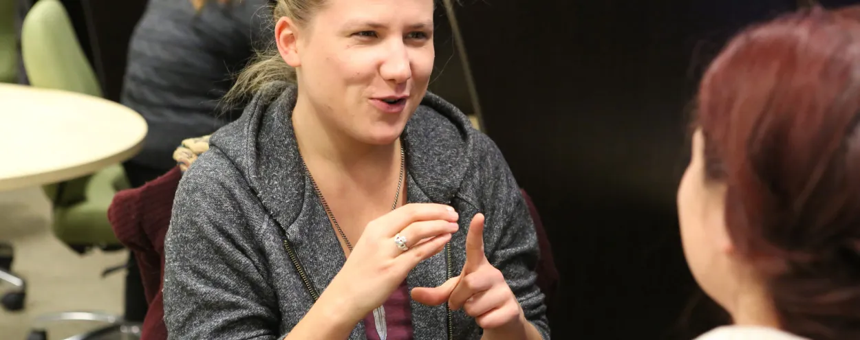 Female American Sign Language and Deaf Studies student practices American Sign Language with another female student seated at video monitor in ASL lab cubicles.