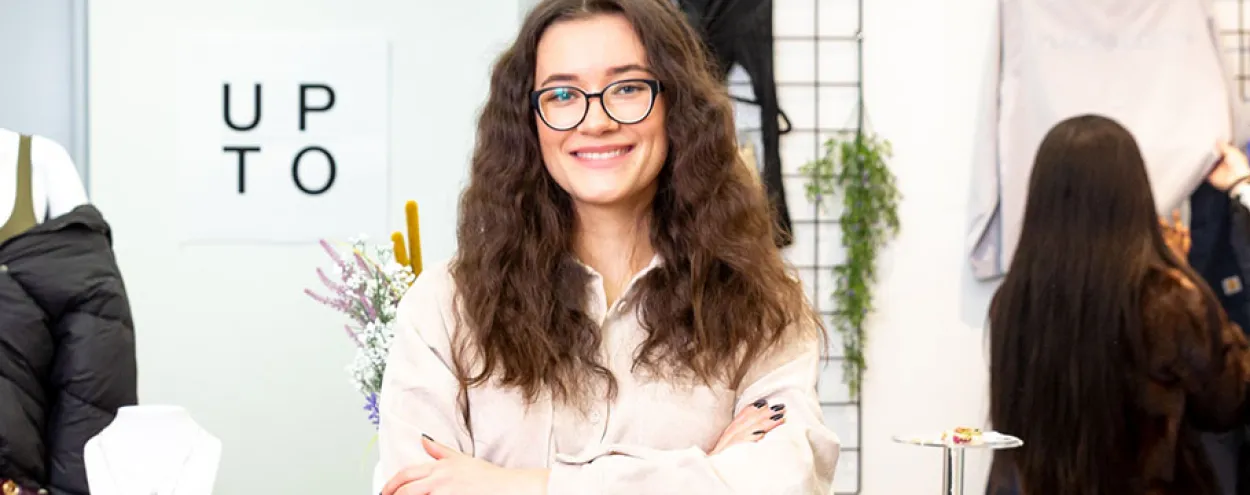 A fashion student stands at a desk and smiles at the camera.