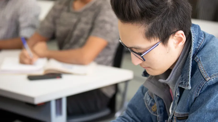 A male student sitting at a desk taking notes during a class.