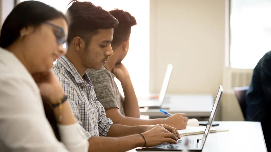 Two male students and one female student taking notes in class with a laptop and paper and pen.
