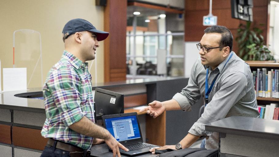 A male student is standing at the library desk and showing settings on his laptop to the librarian while the librarian is pointing at the laptop and explaining something to him.