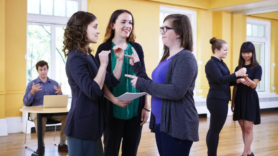 Three female students in the front ground of the photo. Two of the students are practicing sign language while another student observes while holding a piece of paper. In the background there is a male student sitting at a desk in front of a computer. Also in the background, are two female students practicing sign language.