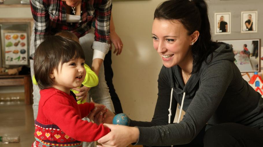 A female child care worker is squatting beside a standing girl toddler. They are both smiling.