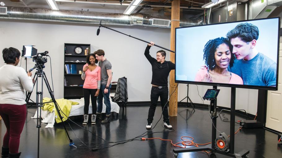 Female and male acting student are in front of the camera in a video studio and being recorded while acting. Closeup of their faces is visible on large TV screen.