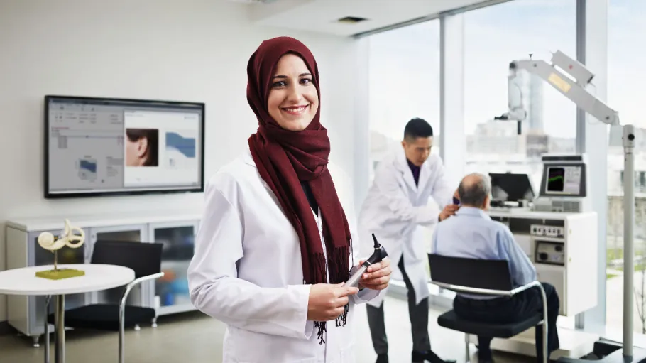 A smiling woman is holding an instrument while standing in the foreground. In the background there is man sitting in a chair who is getting his ear check by another man who is standing.