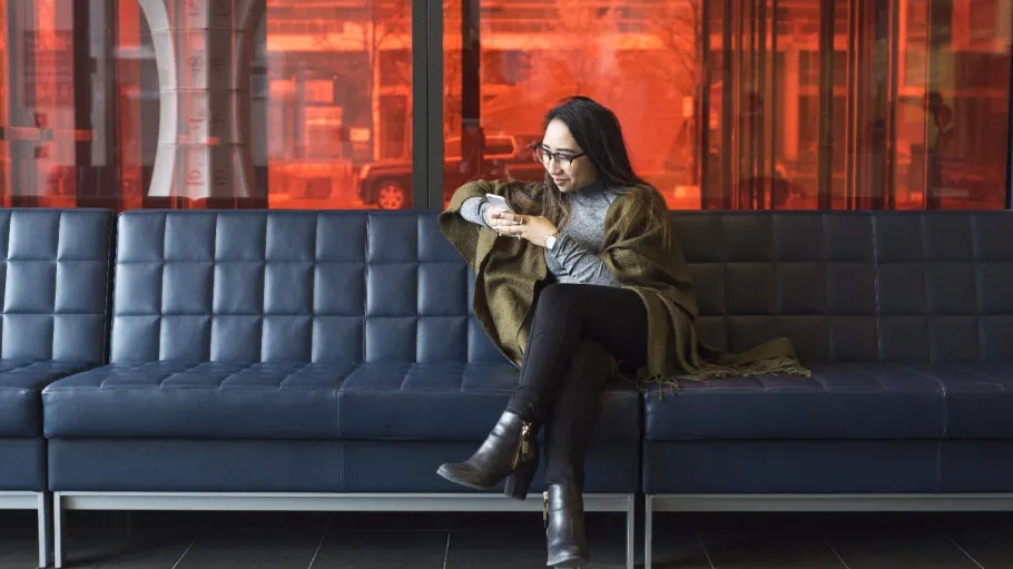 A female student is sitting casually on a comfy lounge bench at the Waterfront Campus and looking at her phone or texting to someone while smiling. 