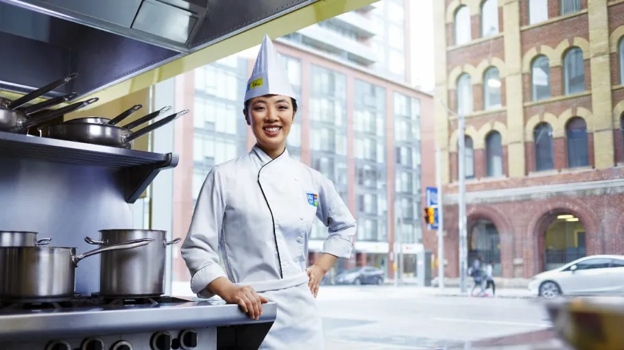 Female Chef School student in uniform poses by stove and window in kitchen/classroom.