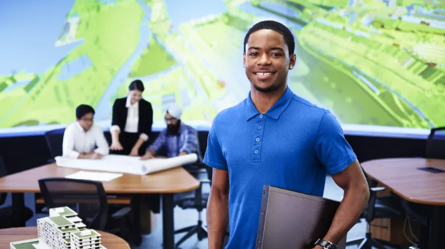 Male architecture student posing with folder in the BIM lab with a 3D model on the table beside him. 