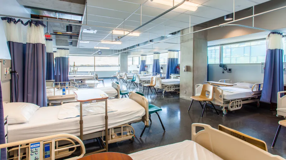 12-bed hospital ward room, Simulation Centre, Daphne Cockwell Centre for Health Sciences. It is a large room with beds. At the end of the beds there are chairs. There are curtains that are around each individual bed: the curtains are drawn back.