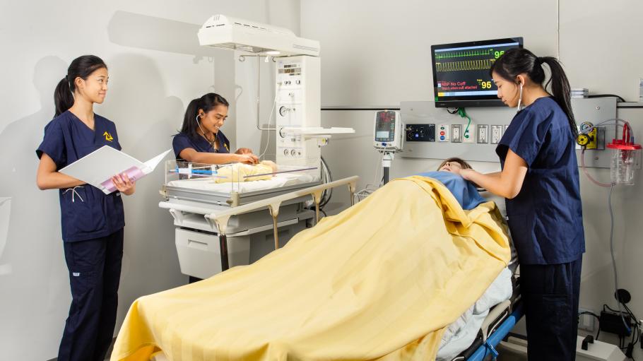 Bachelor of Science in Nursing students practice skills in a high-fidelity suite complete with baby and birthing mother high-fidelity mannequins, Simulation Centre, Daphne Cockwell Centre for Health Sciences. Three female students practicing in the simulation practice centre. One student is standing over a baby dummy measuring its heartbeat. Another student is observing a a screen which has the heartbeat rate. In her hand is a binder. 