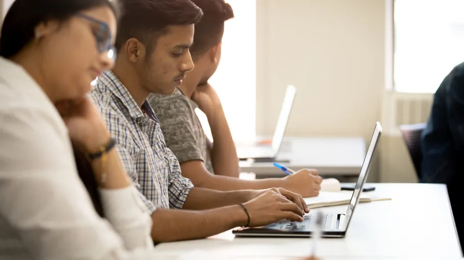 Two male students and one female student taking notes in class with a laptop and paper and pen.