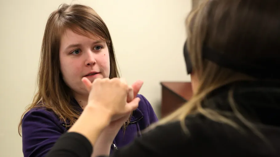 Female students practice two-hand manual communication seated in classroom while one is blindfolded.