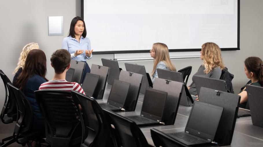 A female instructor in a classroom talking to a group of male and female students sitting in front of laptops.