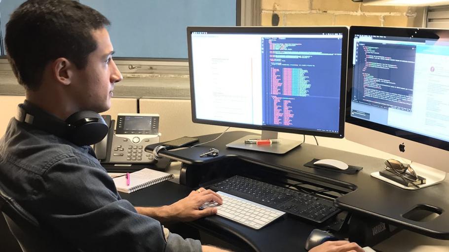 A male employee working at a desk with two computers coding.