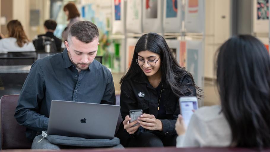 Two students sitting beside each other, one looking at a computer, the other at a phone.