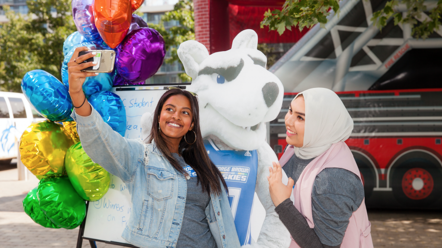 Two students taking a selfie in front of a cutout of the husky mascot.