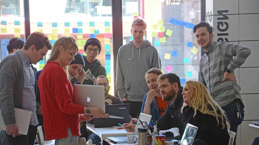 Group of people sitting and standing around a table during a meeting while someone is presenting.