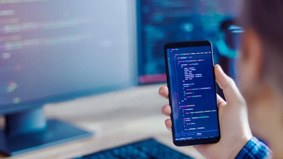 A hand holding a mobile phone with computer code on it in the foreground with a desktop computer in the background.