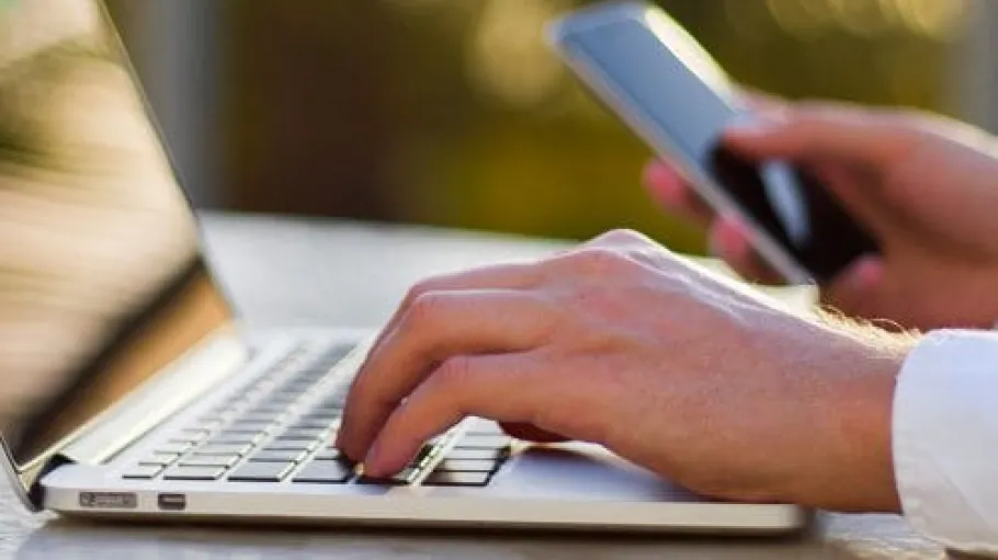 Close up of a hand typing on a laptop and holding a mobile phone with the other hand