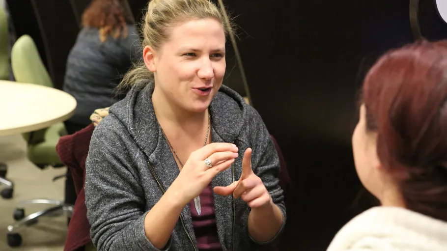 Female American Sign Language and Deaf Studies student practices American Sign Language with another female student seated at video monitor in ASL lab cubicles.