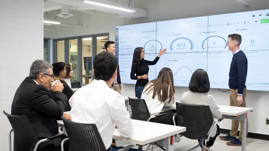 Students present to classmates and faculty on a large digital screen in a brightly lit classroom