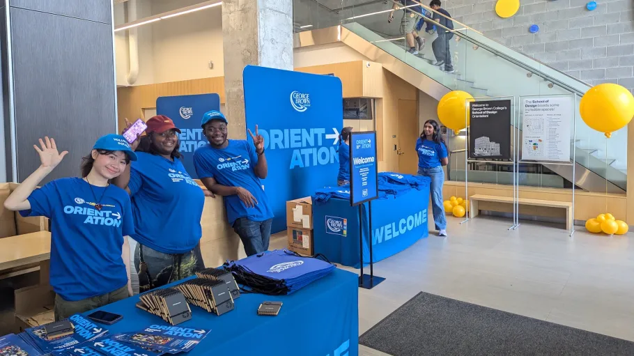 Volunteers at an Orientation Campus Day wave to the camera from behind a table surrounded by balloons.
