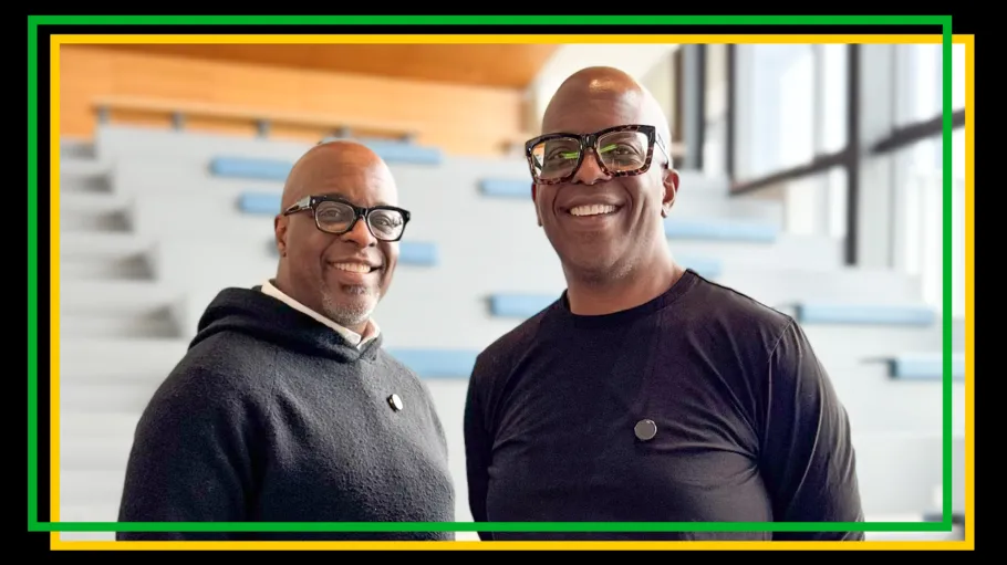 Two men in black shirts and black framed glasses smile