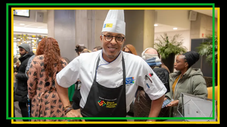 A man in chef whites and hat wearing a black apron smiles