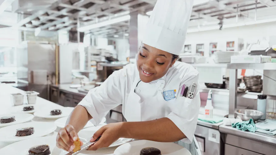 A women in chef whites prepares plates of desserts in a brightly lit kitchen