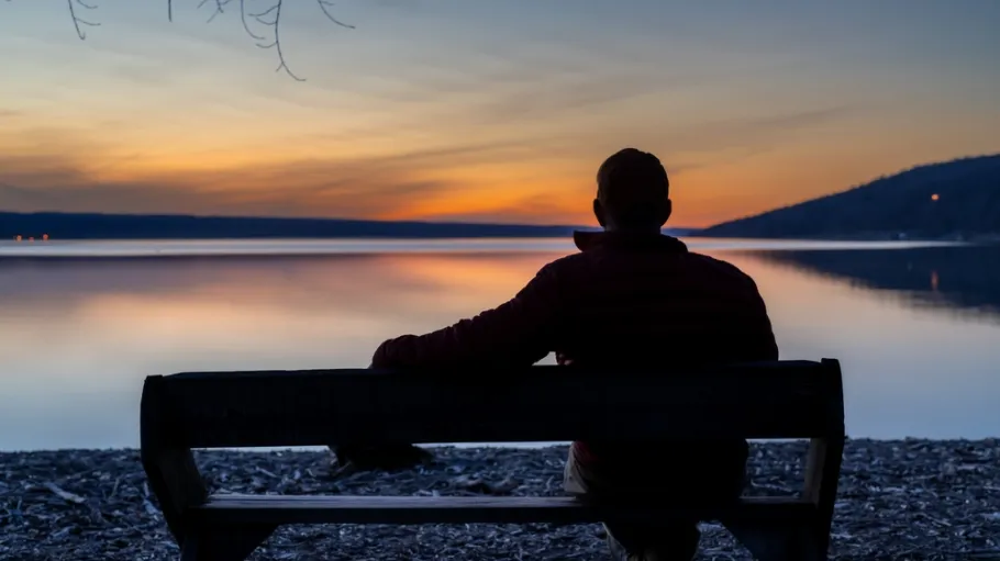 Winter scene of a man with red jacket on a bench looking out over a lake at sunset. Taking time for personal reflection, introspection, thinking about the past or the future. 