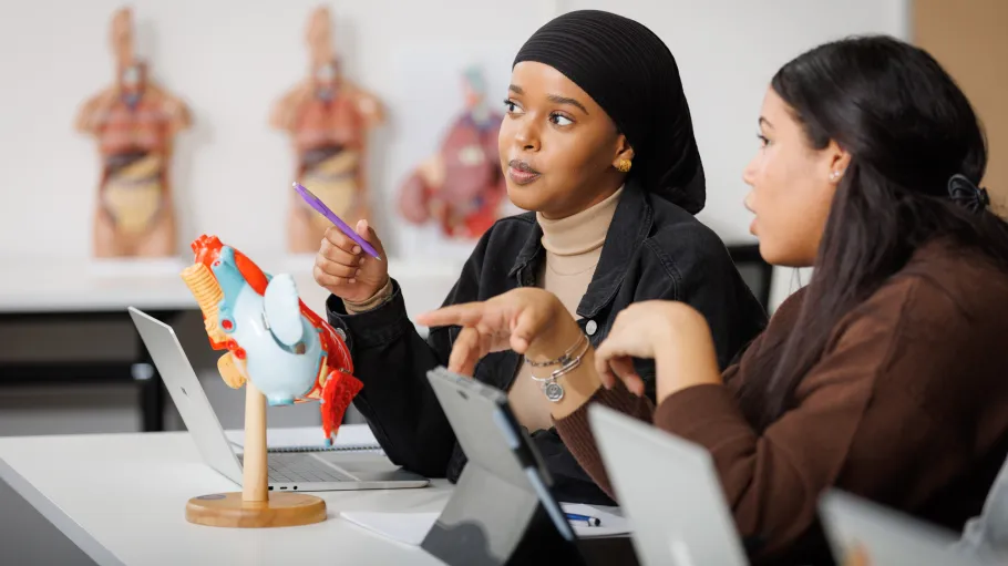 Two students chat at a desk with their laptops open