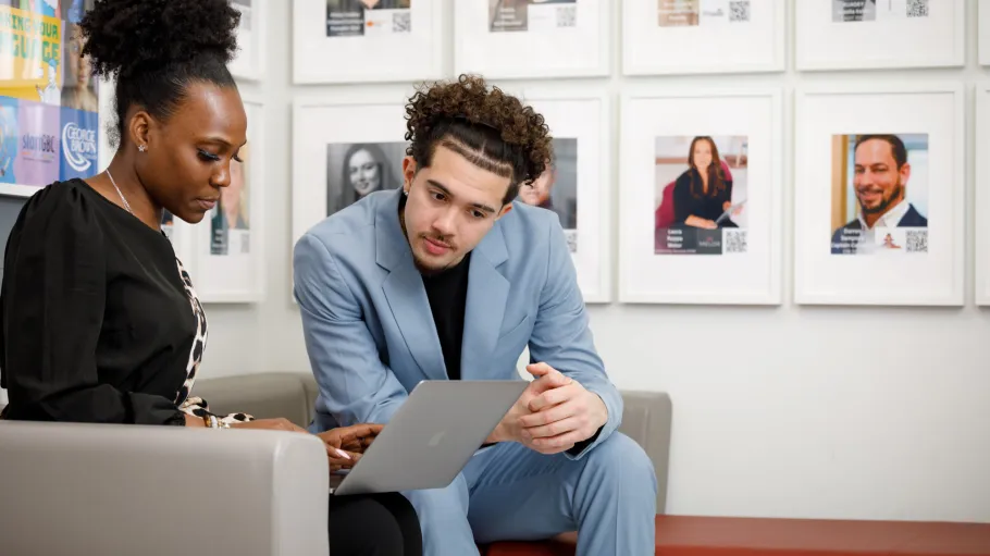 Two seated people looking at a laptop screen in a brightly lit room with pictures on the wall
