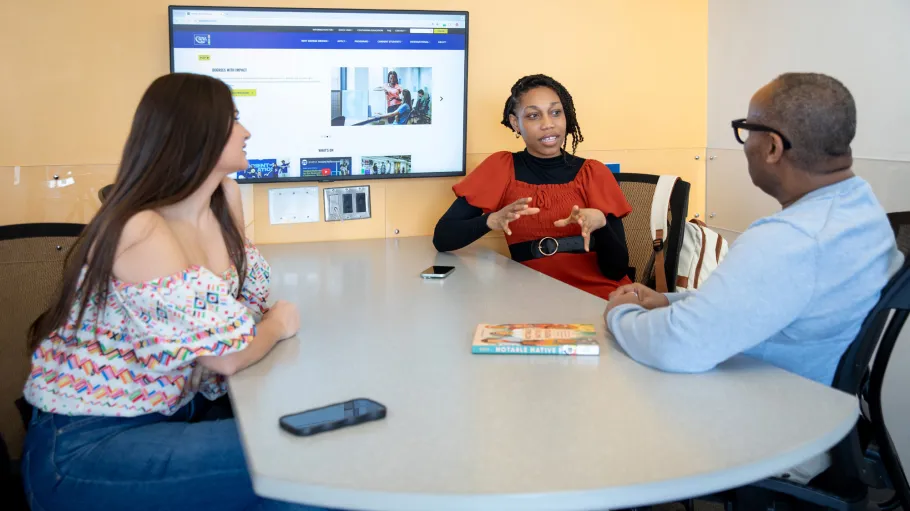 Three students sitting in a Library group study room with a large display screen.