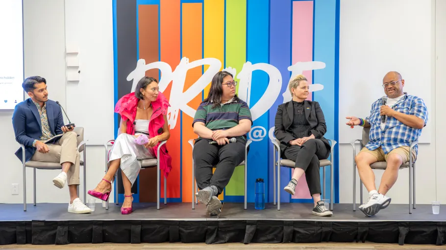 A group of panelists sits on a stage in front of a large colourful background with the word 'Pride'