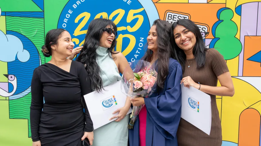 Four GBC graduates pose with their credentials following graduation in front a bright colourful mural