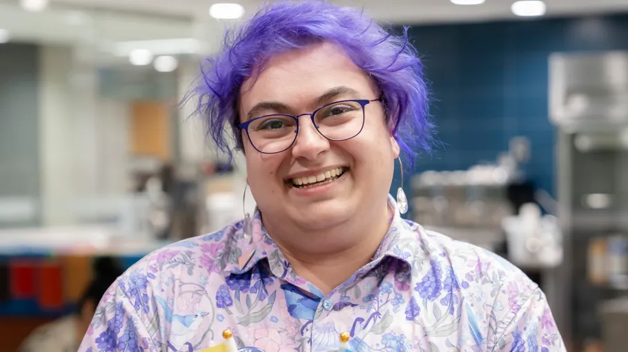 A staff member with purple hair and shirt holds two flags and smiles