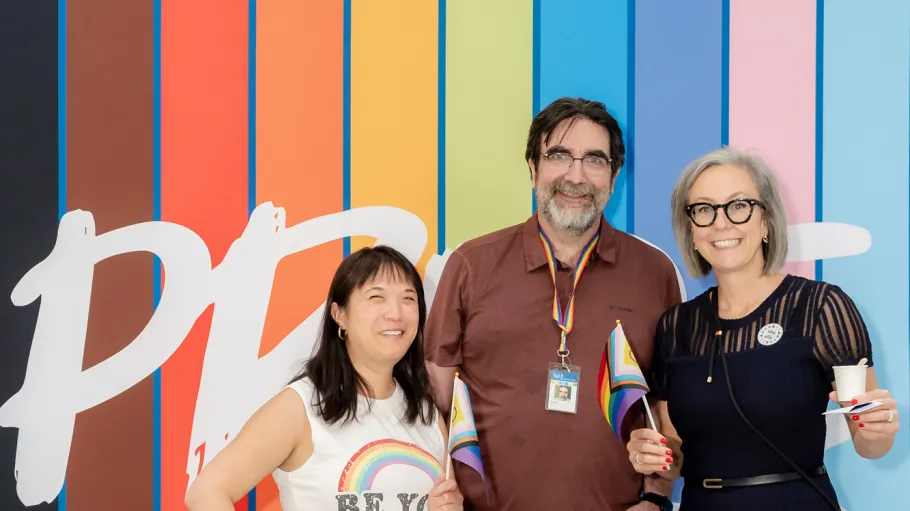 Three community members wave Pride flags in front of a colourful pride banner