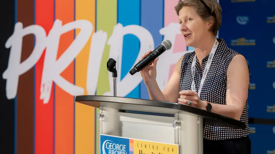 Karine Morin speaks into a microphone at a podium with a colourful pride banner in the background