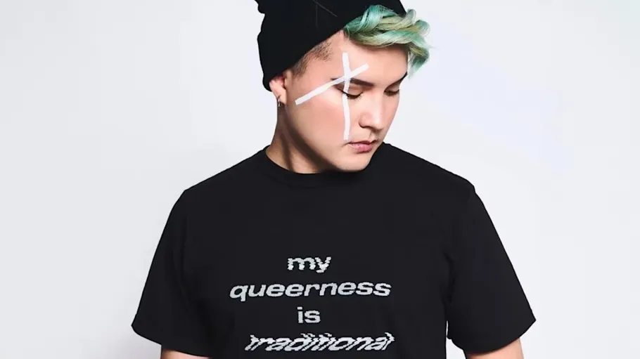 A person with green hair and a black hat and shirt that reads "my queerness is traditional"