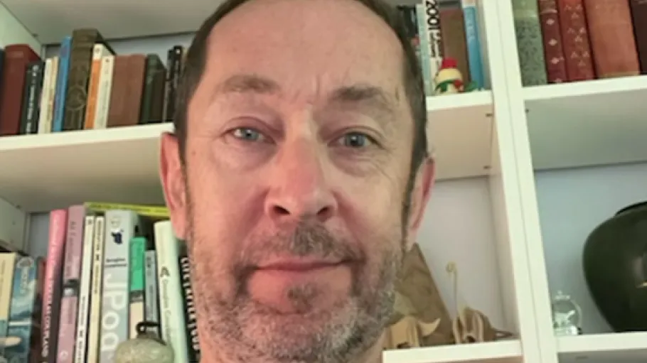 A man in a black t-shirt with facial hair in front of a bookshelf