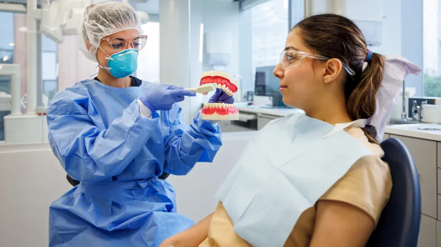 A dental assisting student wearing a light blue gown hairnet and mask holds a model of human teeth to demonstrate how to brush one's teeth to a client sitting in a dental chair.