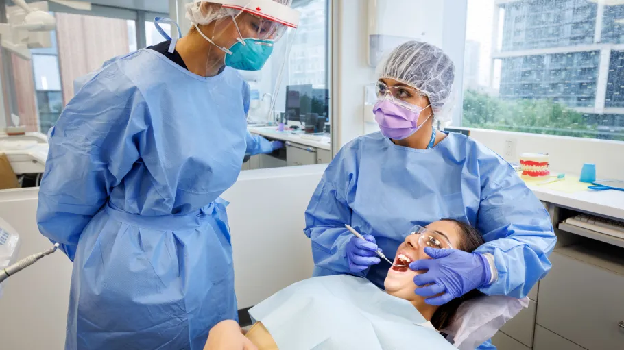 A client reclines in a dunking chair wearing protective eyeglasses as two dental assisting students work on cleaning the client's teeth.