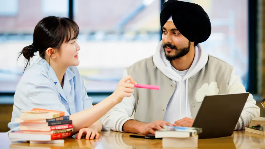 Students sit at a table with an open laptop and discuss the reading materials they have in front of them.