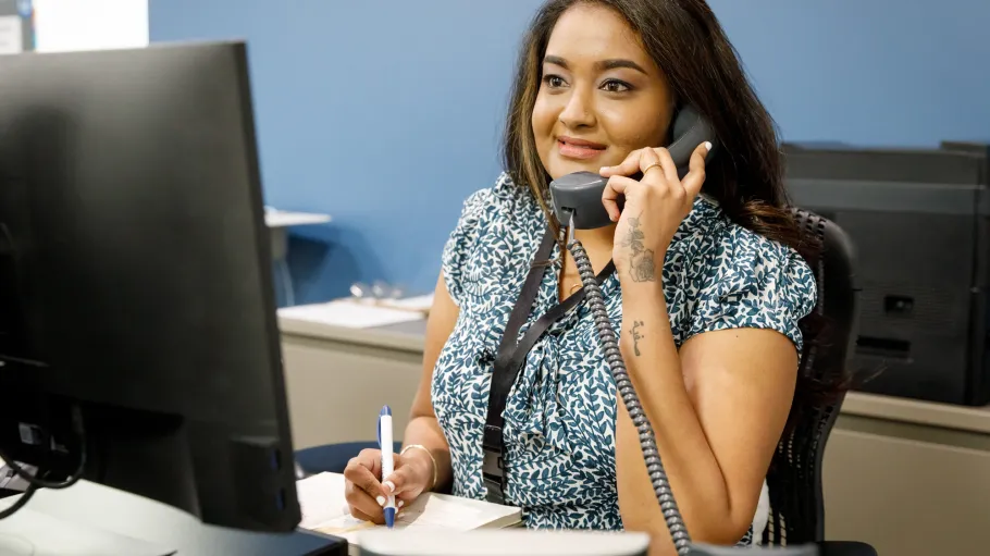 A students sits at a desk in front of a monitor. The student is on the phone with someone and looks at the monitor while taking notes.