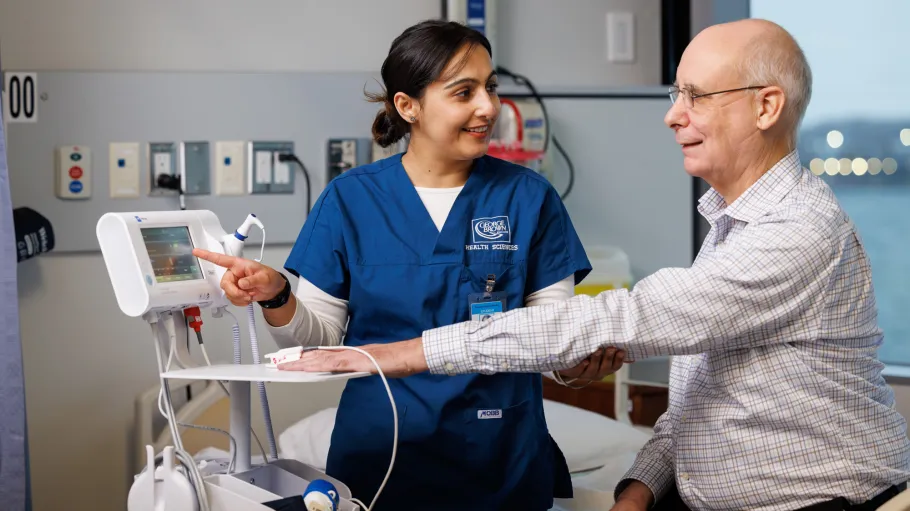 A student in dark blue scrubs measures an elderly client's vitals signs using medical equipment at the Sally Horsfall Eaton School of Nursing.