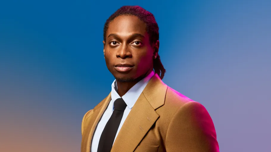 A Black man in a beige suit stands against a colourful blue gradient