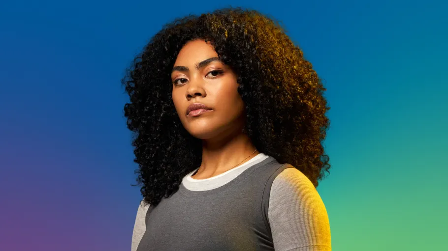 A woman with curly dark hair and grey top stands against a colourful blue gradient