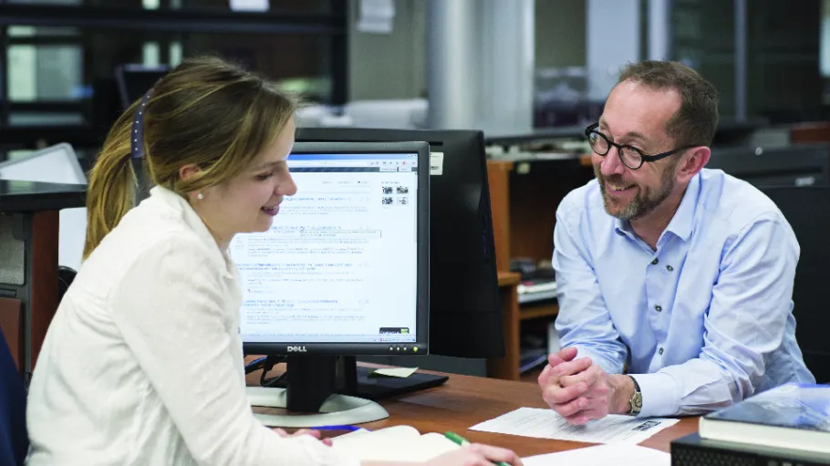 A smiling student sitting with a smiling librarian at the library reference desk looking over an assignment.
