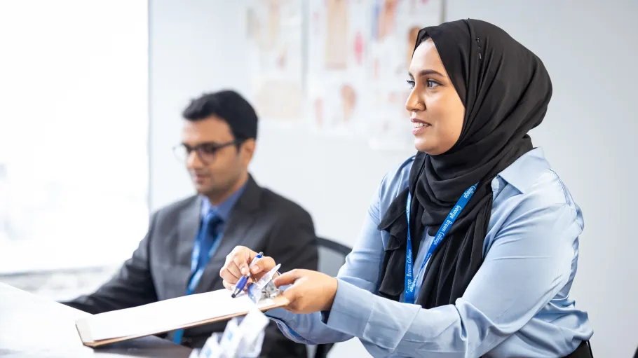 Person at a George Brown Polytechnic reception desk holding a clipboard and pen while assisting a visitor, with another staff member seated nearby.